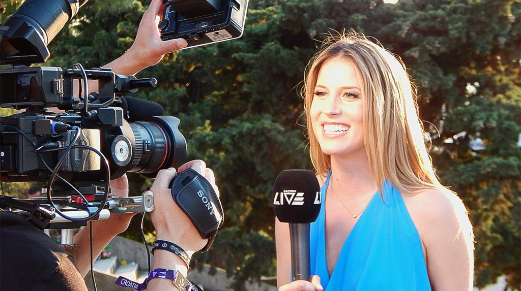A news reporter with a microphone, with a news camera in the left of the shot.