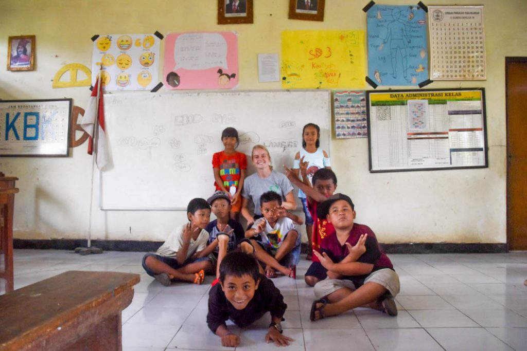 Volunteer teacher Ellen sits with a group of smiling school children on the floor of a classroom.